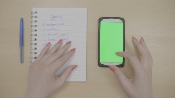 Vue du dessus des mains de la femme avec des ongles rouges touchant smartphone avec écran vert et en regardant la liste de bloc-notes avec des ingrédients de la recette — Video