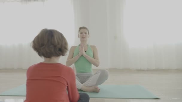 Молодая красивая женщина учит yoga представляет к маленькой девочке ребенка и как медитировать на циновке в студии фитнеса — стоковое видео