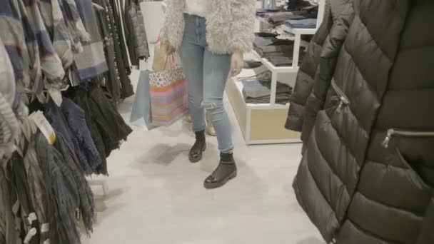 Close up de pernas de mulher vestindo uma roupa casual andando e à procura de roupas com sacos de compras em suas mãos na loja do shopping — Vídeo de Stock