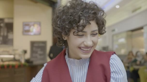 Hermosa hembra con el pelo rizado corto sonriendo y usando su teléfono inteligente mientras bebe de la taza en la cafetería en el centro comercial — Vídeo de stock