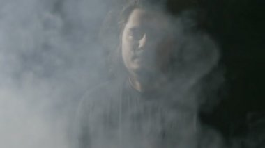 Toplum kurallarına uymayan genç adam uzun saçlı vaping ve Buhar kameraya doğru büyük bulutlar üfleme