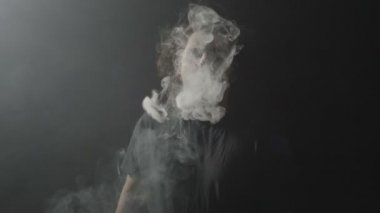 Toplum kurallarına uymayan genç adam vaping elektronik sigara ve etkileyici yapma çevrelerin karanlık bir arka plan üzerinde kameraya doğru duman