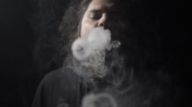 Ağır çekim yetişkin bir adam ayakta bir karanlık odada yalnız vaping elektronik sigara ve duman Buhar daireler kameraya doğru gönderme