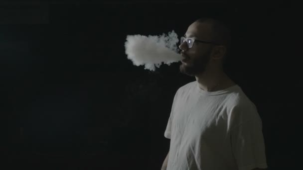 Mladí bokovky chlapec s brýlemi vaping z výparníku a vydechování velké mraky kouře opakovaně v profesionálním studiu tmavě — Stock video