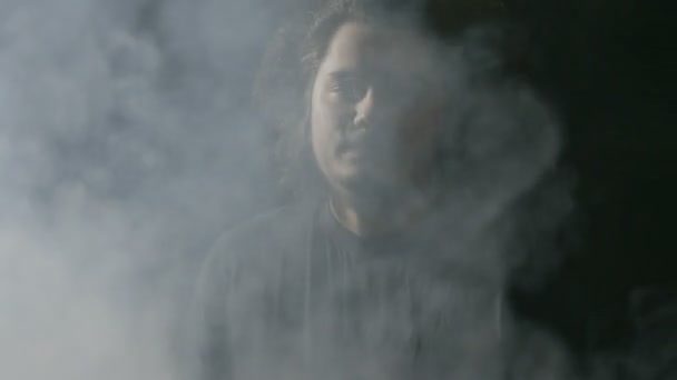 Nekonformní mladý muž s dlouhými vlasy vapingu a foukání obrovská oblaka páry směrem k fotoaparátu — Stock video