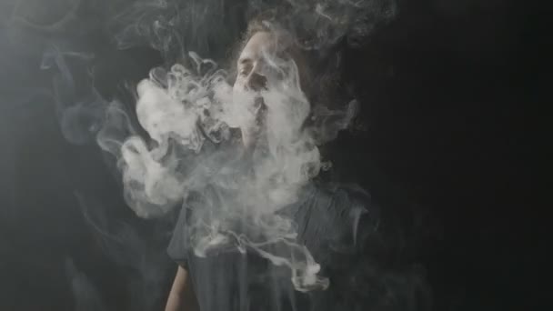 Junger Rocker mit Bart und langen Haaren, der in einem professionellen Studio mit Dampftricks Werbung für elektronische Zigaretten macht — Stockvideo