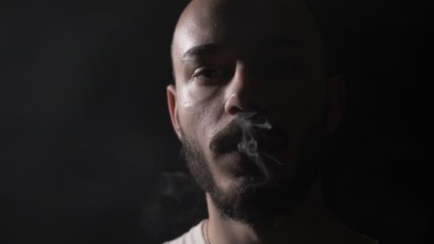 Bold young convict male with beard vaping electronic cigarette and exhaling a large cloud of smoke towards camera in a dark cold room of prison — Stock Video