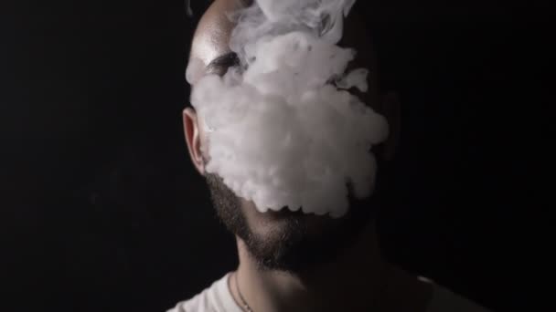 Bold young male musician vaper with beard vapes and blows on his mouth a huge cloud of vape smoke that covers his face in slow motion — Stock Video