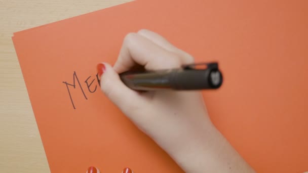 Mulher mãos escrevendo Feliz Natal em letras maiúsculas em um papel vermelho com um marcador preto — Vídeo de Stock