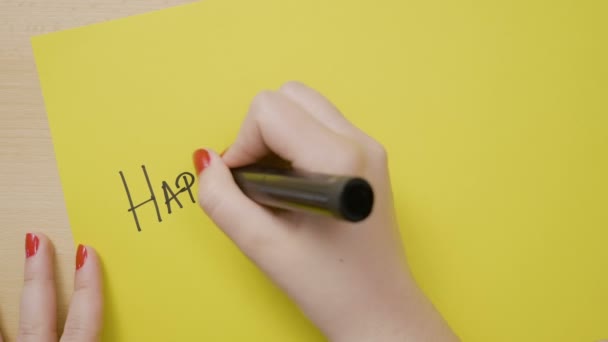 Руки молодої студентки з червоними цвяхами пишуть щасливі свята в великих листах на жовтому папері з чорним маркером — стокове відео