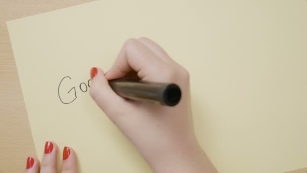Giovani mani femminili che scrivono buongiorno e disegnano una faccina sorridente su una carta gialla con un pennarello nero — Video Stock