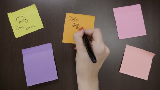 Vista superior de las manos de la mujer escribiendo en el poste pegajoso de color que notas citas motivacionales utilizando pluma negra — Vídeo de stock