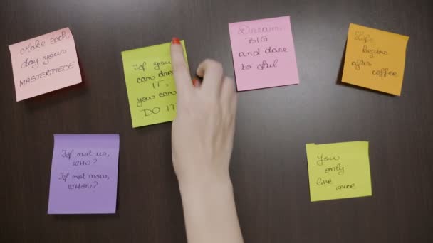 Jonge tiener meisje met rode nagels steken twee post-it notities waarin motiverende citaten naast anderen op een donkere houten bureau — Stockvideo