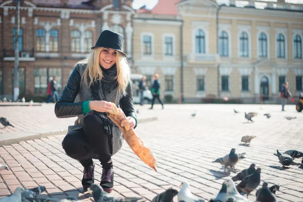 Young beautiful woman is feeding pigeons with bread crumbs in the town square — Stock Photo, Image