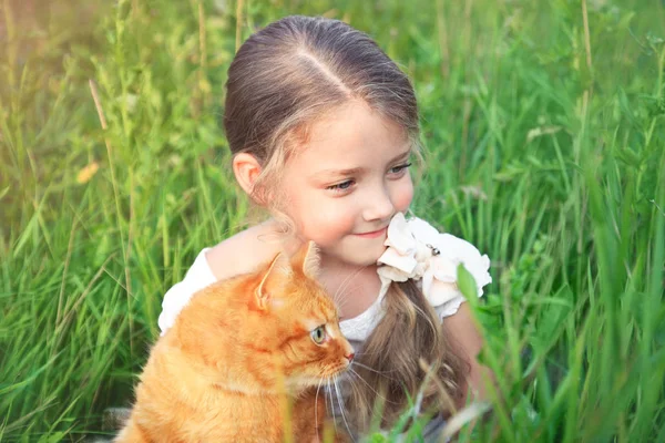 かわいい女の子は、草の中に座って赤猫を保持しています。. — ストック写真