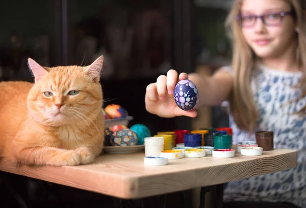 Genç kız Paskalya yumurtalarını boyar, kızıl kedisi masanın üstünde yatar.. — Stok fotoğraf