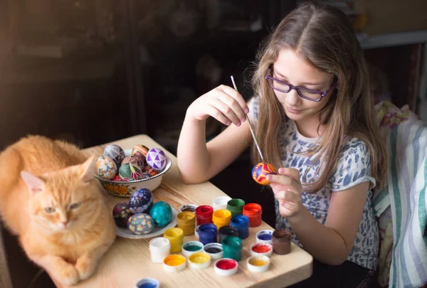 Teen girl paints Easter eggs, her ginger cat lies on the table.