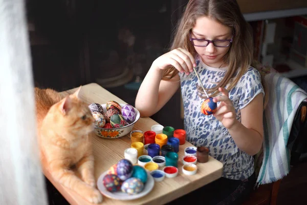Teenie-Mädchen bemalt Ostereier, ihre Ingwerkatze liegt auf dem Tisch. — Stockfoto