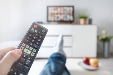 Televizyon izleyen bir adam, kanepeye uzanmış, bacakları masanın üstünde. Oturma odasında uzaktan kumandalı biri var.