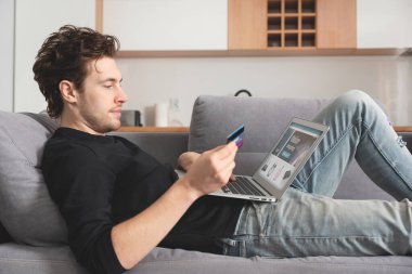 Kredi kartıyla genç alışverişi. Adam dizüstü bilgisayarla kanepede yatıyor.