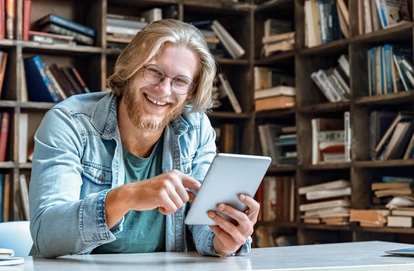 Young bearded man glasses library desk hold modern tablet laugh at camera.