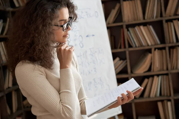 Young latin math school teacher wearing glasses holding notebook in classroom.