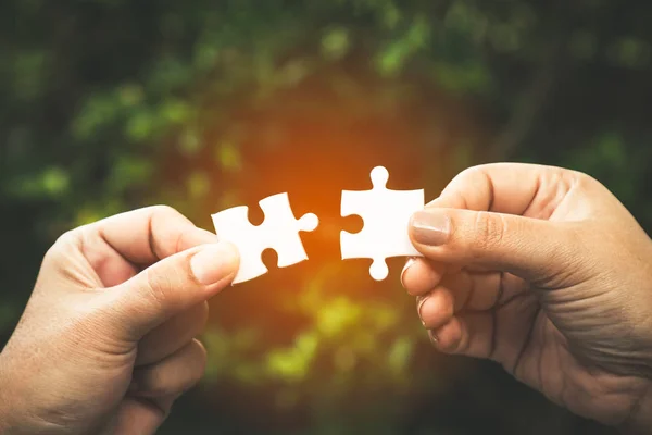 two hands trying to connect couple puzzle piece with forest background; one part of whole; symbol of association and connection for business strategy.