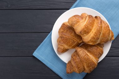 Beyaz tabakta kruvasan ve siyah tahta bir masa. Mavi peçeteyle servis edilmiş. Taze Fransız kruvasanı. Sıcak taze tereyağlı rulo. Metin için boş alan. Yukarıdan bak. Üst görünüm.