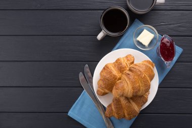 Siyah ahşap bir masada taze kruvasanlar. Kahve, kahve demliği, tereyağı, reçel ve mavi peçeteyle servis edilir. Taze Fransız kruvasanı. Sıcak taze tereyağlı rulo. Kahvaltıda. Metin için boş alan. Yukarıdan bak. Üst görünüm. 