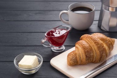 Siyah ahşap bir masada taze kruvasanlar. Kahve, kahve demliği, tereyağı, reçel ve mavi peçeteyle servis edilir. Taze Fransız kruvasanı. Sıcak taze tereyağlı rulo. Kahvaltıda. Metin için boş alan. 