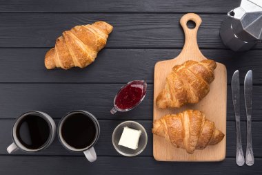 Siyah ahşap bir masada taze kruvasanlar. Kahve, kahve demliği, tereyağı, reçel ve mavi peçeteyle servis edilir. Taze Fransız kruvasanı. Sıcak taze tereyağlı rulo. Kahvaltıda. Metin için boş alan. Yukarıdan bak. Üst görünüm. 