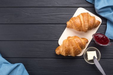 Siyah ahşap bir masada taze kruvasanlar. Kahve, kahve demliği, tereyağı, reçel ve mavi peçeteyle servis edilir. Taze Fransız kruvasanı. Sıcak taze tereyağlı rulo. Kahvaltıda. Metin için boş alan. Yukarıdan bak. Üst görünüm. 