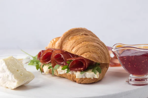 Fresh croissant sandwich with ham, cheese and arugula on white wooden table. Served with Coffee, cheese, ham, orange juice, jam. Fresh French Baked Croissants. Free space for text.