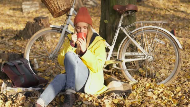 快乐而微笑的女人坐在靠近树和自行车的地面上 用红杯喝茶 她很快乐 微笑着 享受着美好的秋日 — 图库视频影像