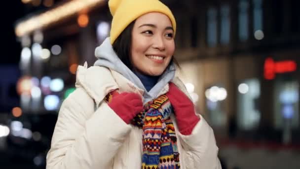 笑顔とかなりアジアの女性が居心地の良い感じを見て 夜の通りで自分自身を抱擁 手持ち撮影 — ストック動画