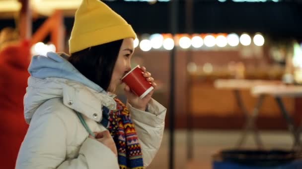 若いアジア人女性が冬休みに通りを歩いている 熱いお茶と笑顔を飲む 手持ち撮影 — ストック動画