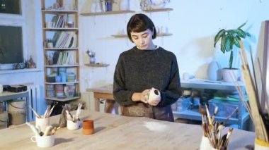 Çömlekçi kadın seramik bardağını tutuyor ve öğütüyor. Sanat atölyesinde durmuş, kameraya bakıyor. Yavaş çekim.