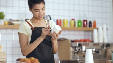 Gülümseyen ve arkadaş canlısı görünen Afro-Amerikan kahveci elinde kağıt bardak ve üzerine müşterinin adını yazıyor. Kahve makinesine yürüdükten sonra. El kamerasıyla..