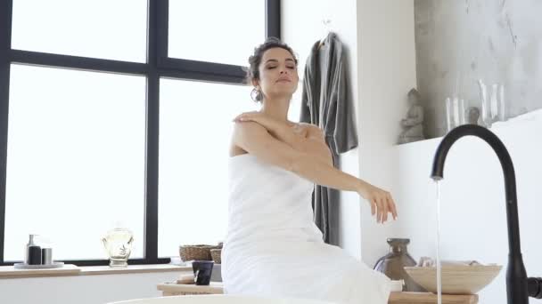 浴槽に座っているタオルで服を着たブルネットの女性は 手と首にクリームを適用します スキンケア スローモーション — ストック動画