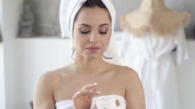 Banyo sonrası cilt bakımı. Yüzüne krem süren genç bir kadının portresi. El kamerasıyla..
