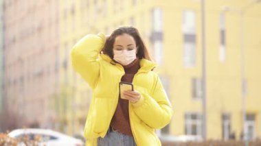 Yüz maskesi takan, cep telefonu tutan ve sokakta yürüyen bir kadın. El kamerasıyla..