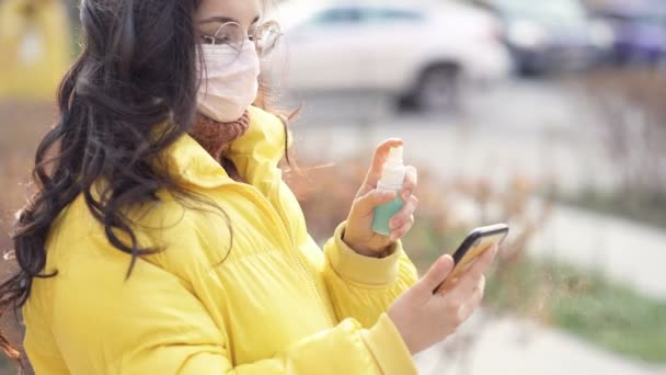 在智能手机上使用清洁剂喷雾的戴口罩的年轻女子 — 图库视频影像