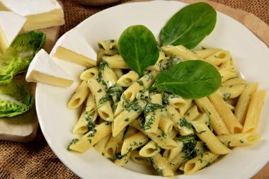 Beyaz tabakta ıspanak soslu makarna yanında taze ıspanak yaprakları ve ahşap mutfak tahtasındaki mavi peynir.