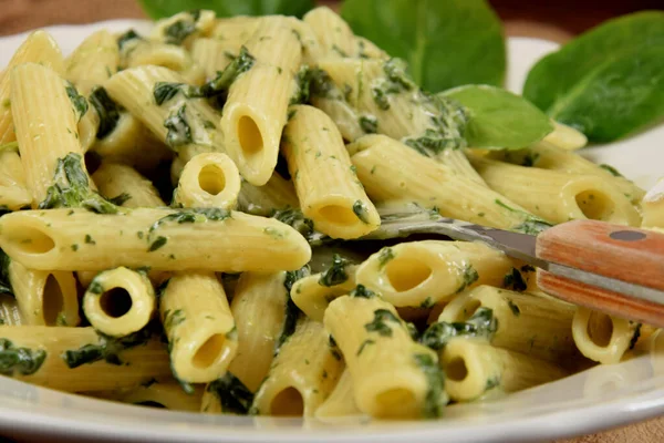 penne in spinach sauce on a white plate in the company of fresh spinach leaves and blue cheese on a wooden kitchen board