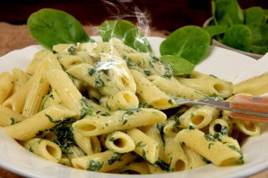 Beyaz tabakta ıspanak soslu makarna yanında taze ıspanak yaprakları ve ahşap mutfak tahtasındaki mavi peynir.