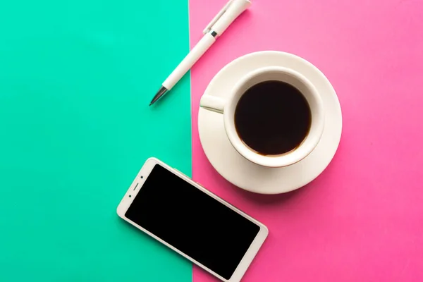 Flat lay photo with coffee cup, mobile phone and pen on geometric green and pink background. Top view, minimal concept