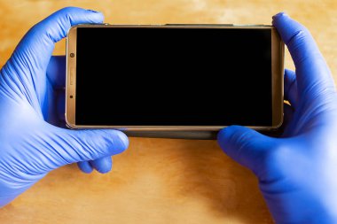 Tıbbi eldivenlerdeki eller ahşap arka planda cep telefonunu tutuyor.