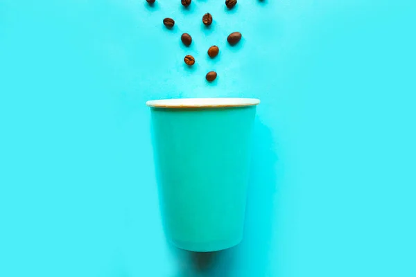 Empty take away coffee cup and coffee beans on blue background. Minimal concept