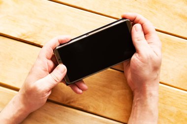 Adam ahşap arka planda siyah ekran model telefonu tutuyor, üst görünümde.