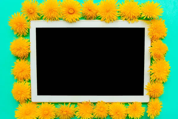 Tablet with black mockup screen with fresh yellow dandelions frame on green background. Copy space for the text. Springtime concept